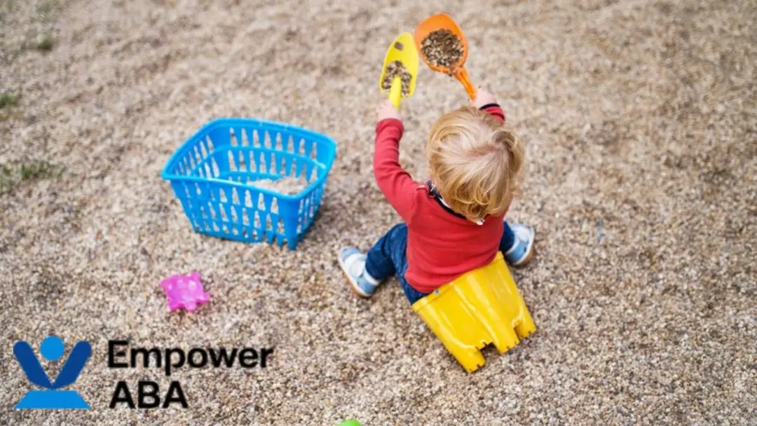 Sensory integration therapy autism: A toddler plays in a sandbox, proudly holding a small plastic shovel filled with sand, exploring texture through play.