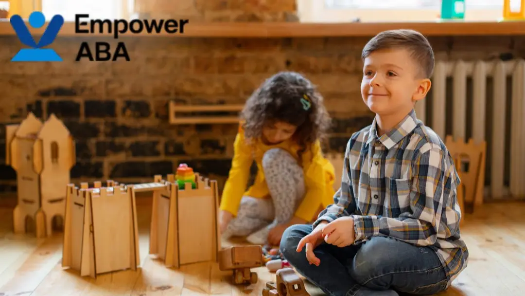 Play schemas autism, a young boy sitting on the playroom floor looks up and smiles, surrounded by toys and other children.