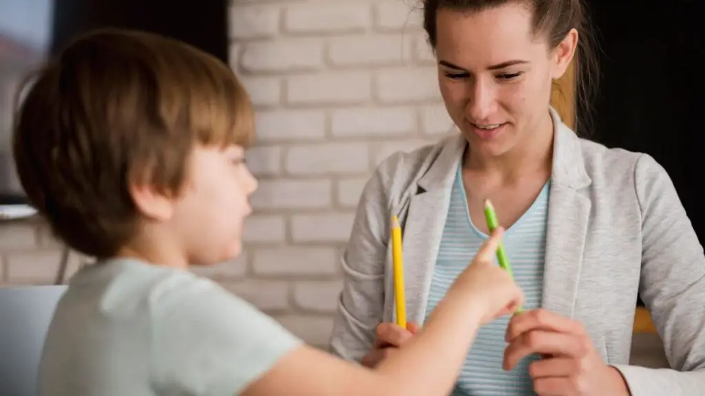In a therapy room, a child therapist gently guides a young child through a social skills task at a table. The therapist and child sit together, focusing on the activity.