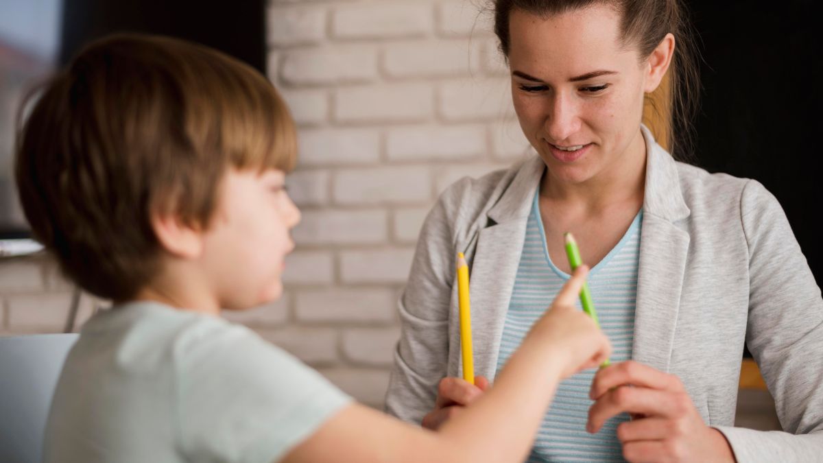 In a therapy room, a child therapist gently guides a young child through a social skills task at a table. The therapist and child sit together, focusing on the activity.
